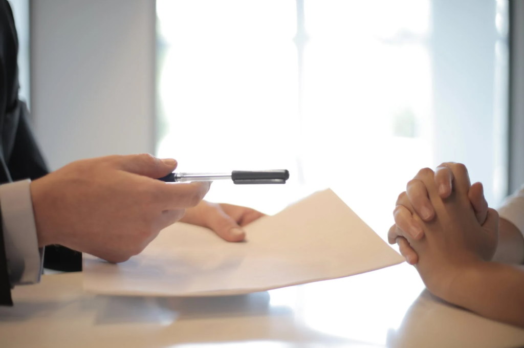 Image of a hand offering a contract and a pen to sign it to a pair of folded hands.