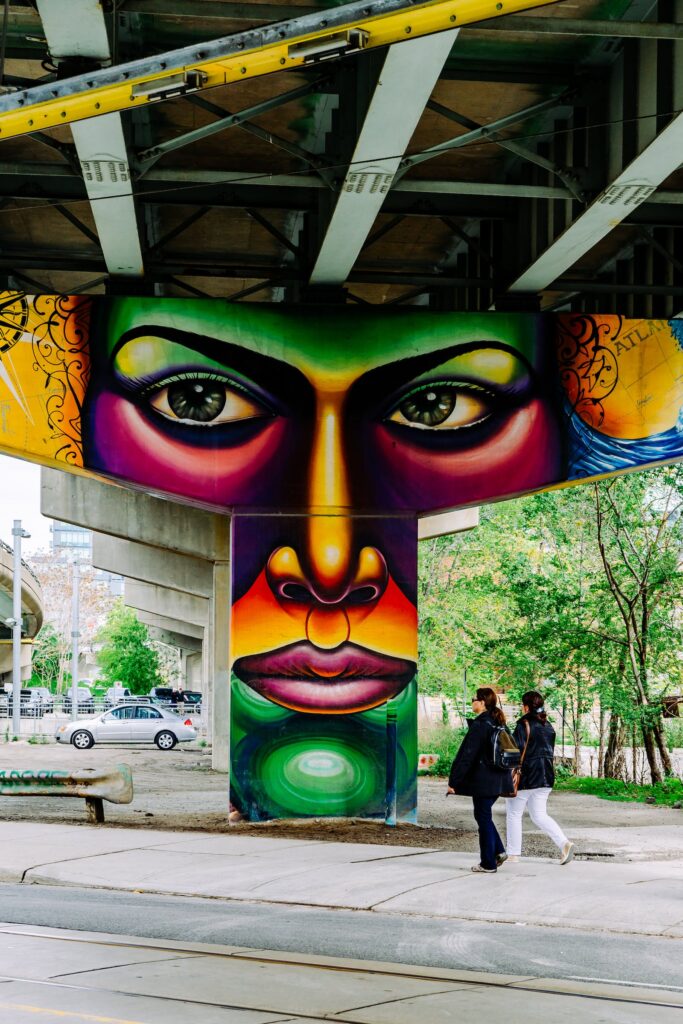 Image of a concrete column under a bridge painted by a street artist depicting a face in a burst of vibrant colors.